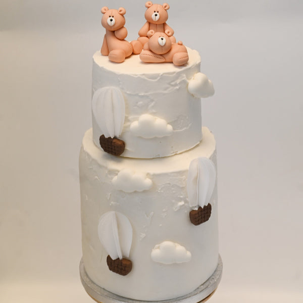 Two-tier white cake decorated with edible clouds, hot air balloons, and three fondant teddy bears sitting on top.