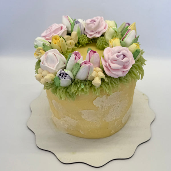 Pastel flower wreath cake with tulips, roses, and buttercream greenery, arranged in a ring on top of a rustic-finish yellow cake.