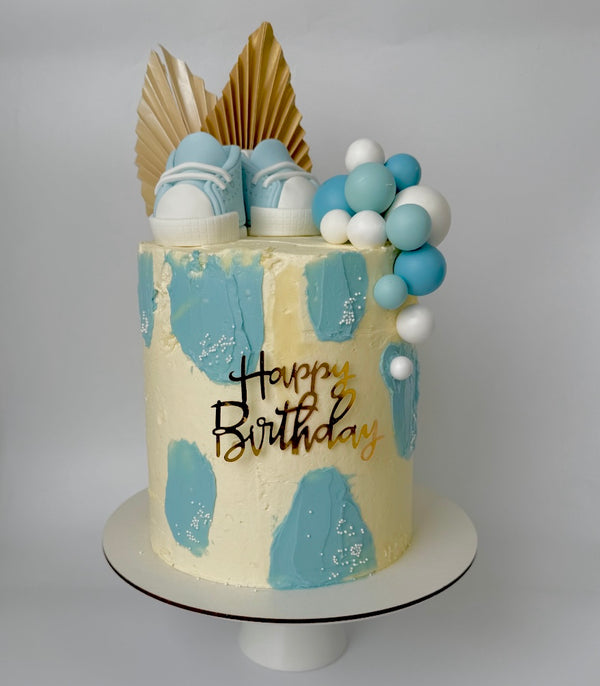 “Blue and beige birthday cake with baby sneakers on top, fondant balloon cluster, golden fans, and a ‘Happy Birthday’ topper.