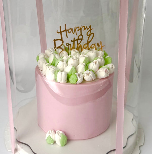 Pink birthday cake designed as a flower box with white and green tulip-shaped decorations and a ‘Happy Birthday’ topper, placed inside a transparent gift box.