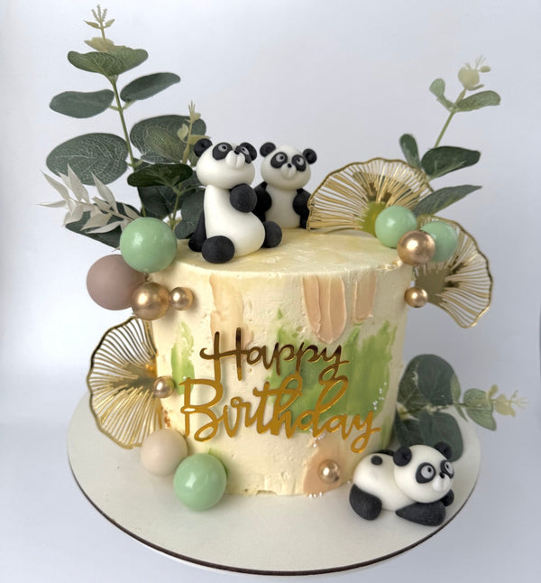 “Jungle-themed birthday cake with panda figurines, green and beige frosting, gold accents, greenery, and a ‘Happy Birthday’ topper.”