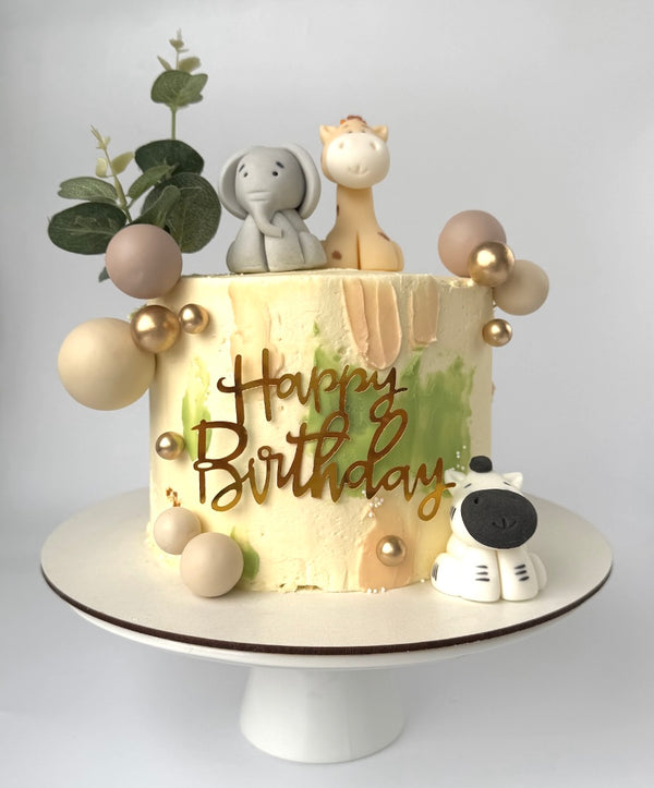 Birthday cake decorated with a giraffe, elephant, and zebra figurines, green and beige frosting, golden edible balls, and a ‘Happy Birthday’ topper.
