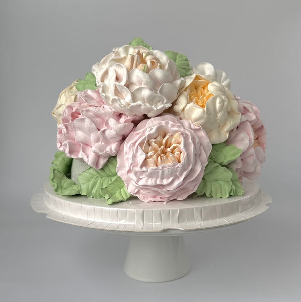 “Elegant floral cake covered with large buttercream roses and peonies in pastel shades of pink, cream, and yellow with green leaves.”