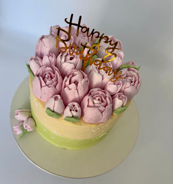 “Floral birthday cake with pink tulip-like buttercream flowers and green accents, featuring a ‘Happy Birthday’ topper.”