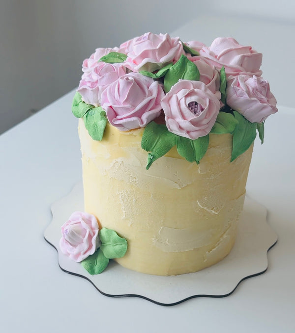 Buttercream birthday cake decorated with pink roses and green leaves on a pale yellow base. Elegant floral design.