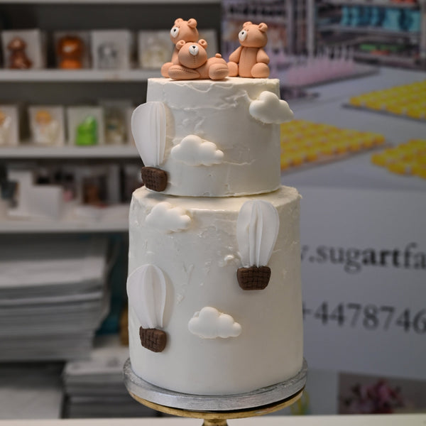 White two-tier cake with fondant teddy bears, clouds, and hot air balloons, displayed at a baking event or pastry shop.