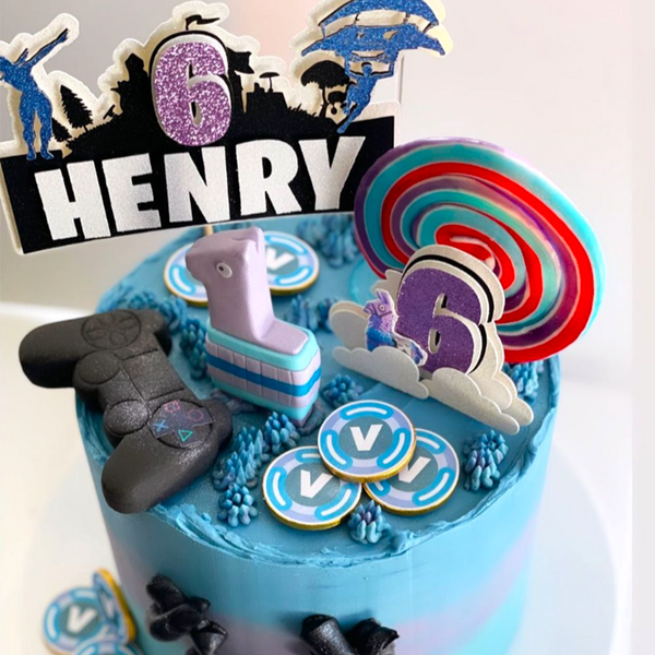 Custom Fortnite-themed birthday cake with edible decorations including a game controller, llama piñata, V-Bucks coins, and a topper reading “Henry 6”.
