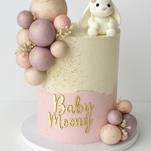 Charming pink and beige baby shower cake with speckled finish, pastel balloon clusters, and a cute fondant bunny topper, labeled ‘Baby Moony’.”