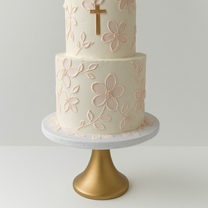 Two-tier white cake with light pink floral decorations and a gold cross on top, displayed on a golden cake stand.