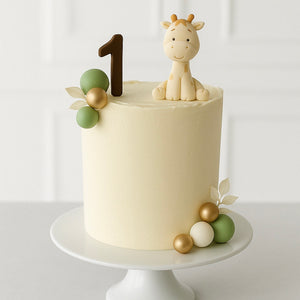 Simple ivory cake topped with a fondant giraffe figure, green and gold edible balls, and a brown number “1” topper.