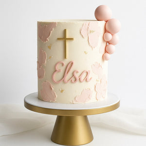 Minimalist cream cake with textured pink accents, a gold cross, and soft pink edible spheres, featuring the name “Elsa” in pink script.