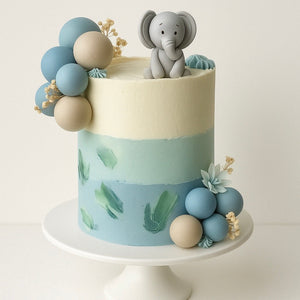 Elegant pastel blue and cream birthday cake decorated with balloons, delicate flowers, and a cute gray elephant edible topper, displayed on a white cake stand.