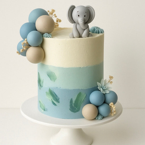 Elegant pastel blue and cream birthday cake decorated with balloons, delicate flowers, and a cute gray elephant edible topper, displayed on a white cake stand.