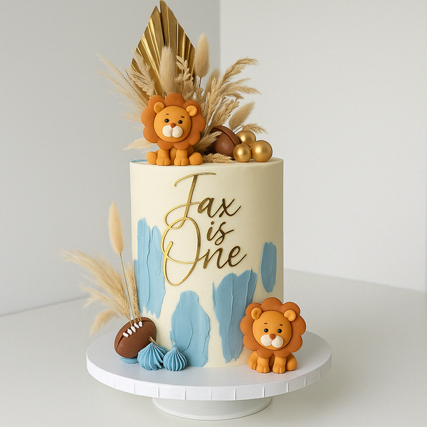 Safari-themed first birthday cake with golden palm leaves, fondant lions, football, and pampas grass, decorated with blue brush strokes and ‘Jax is One’ in gold.