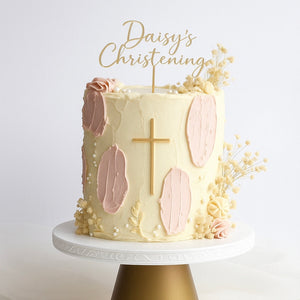 Ivory buttercream cake with blush pink accents, a gold cross, white pearls, and a topper that says “Daisy’s Christening,” finished with dried flowers.