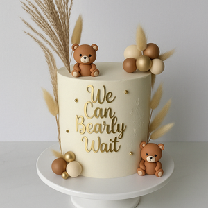 	Beige cake with teddy bears, golden and brown spheres, dried grass, and the phrase ‘We Can Bearly Wait’ in gold letters.