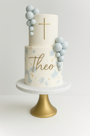 Elegant two-tier cake with gold cross, name "Theo", and clusters of pastel blue fondant balls.