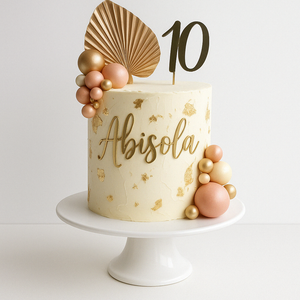 Elegant white cake with gold leaf accents, personalized with the name “Abisola” and a large gold number 10 topper, decorated with pink and gold edible spheres and a golden palm leaf.