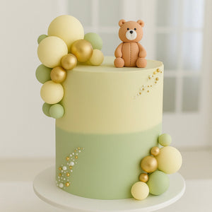 Pastel yellow and green cake with gold and green fondant balls and a small teddy bear on top, placed on a white stand.