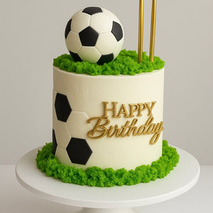 Soccer-themed birthday cake with black and white hexagon pattern, fondant soccer ball on green ‘grass’ topping, and ‘Happy Birthday’ in gold.”