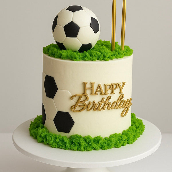 Soccer-themed birthday cake with black and white hexagon pattern, fondant soccer ball on green ‘grass’ topping, and ‘Happy Birthday’ in gold.”