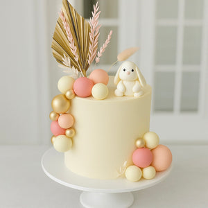 Cream-colored cake with peach and gold balls, a bunny figurine, and dried palm leaves, ideal for baby showers.