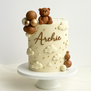 “Neutral-toned baby cake with white and gold clouds, stars, fondant teddy bear, and the name ‘Archie’ in gold script.”