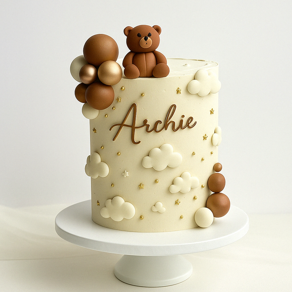 “Neutral-toned baby cake with white and gold clouds, stars, fondant teddy bear, and the name ‘Archie’ in gold script.”