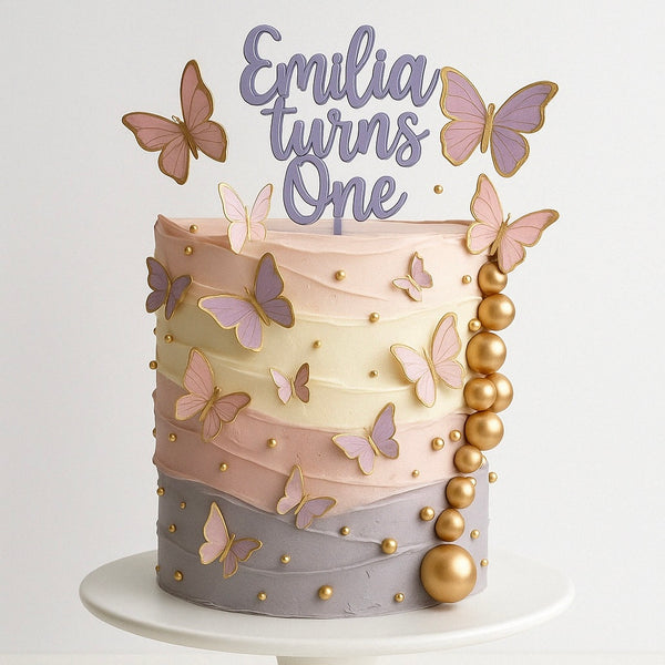 Pastel rainbow-layered cake with purple, pink, and gold butterflies, gold pearls, and a topper reading “Emilia turns One” in lavender letters.
