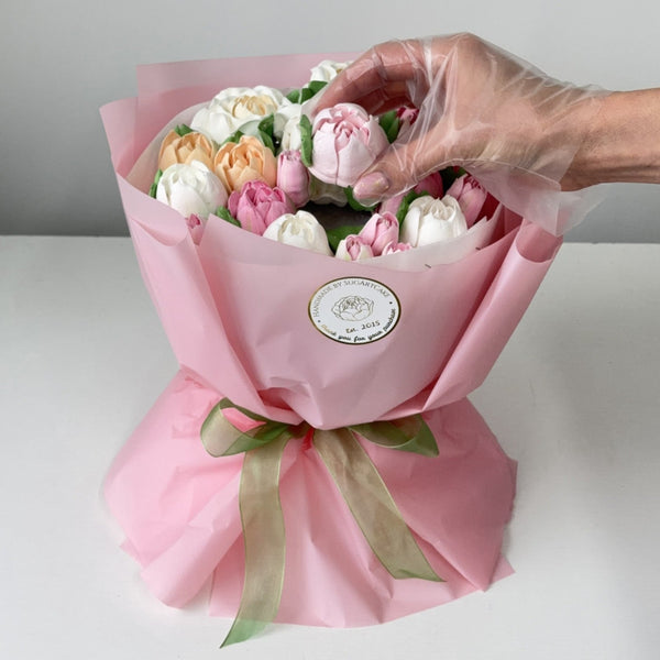 Bouquet of marshmallow tulip flowers wrapped in pink paper with a hand adjusting the flowers, on a light background.