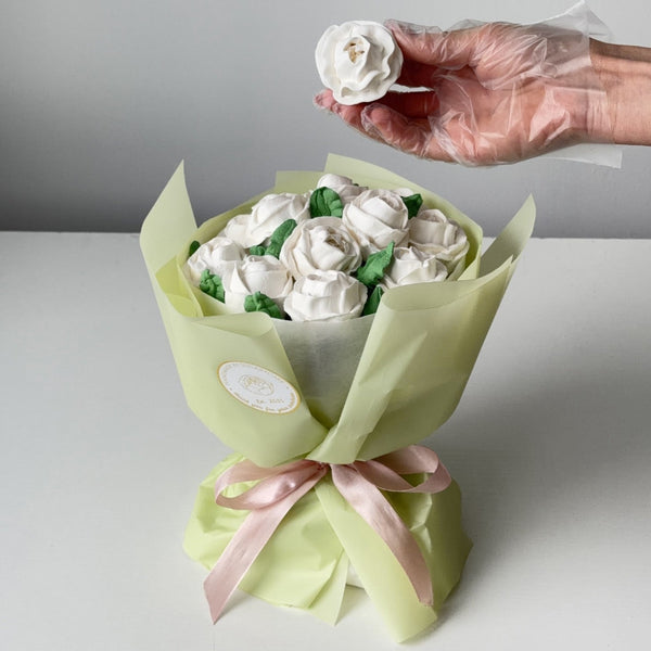 Bouquet of white roses wrapped in green paper with a pink ribbon, held by a gloved hand against a gray background.