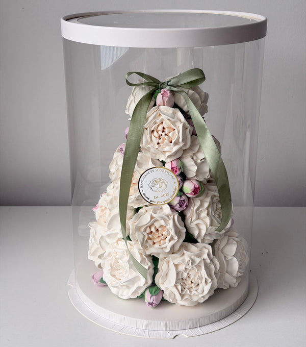 Decorative cake with white flowers and a green ribbon inside a clear dome on a white surface.