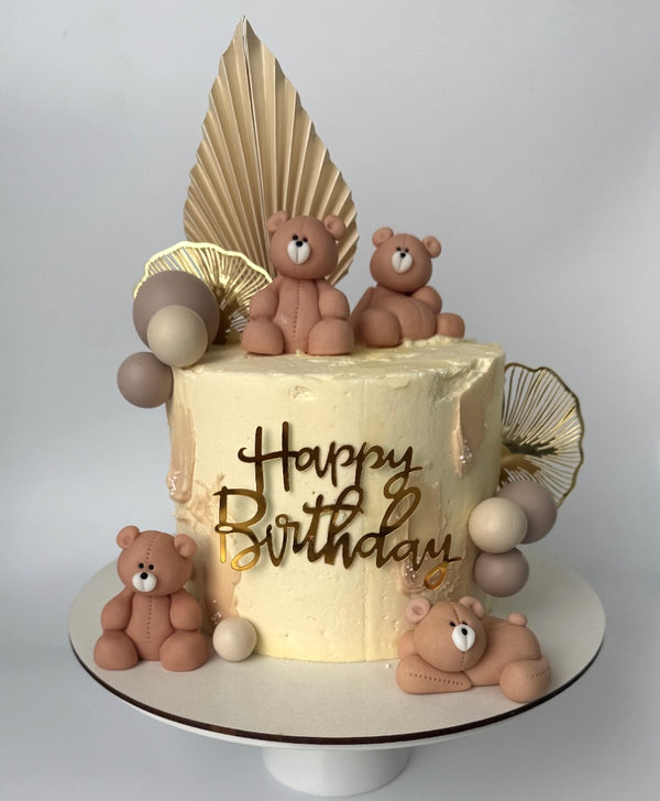 “Birthday cake with teddy bear figurines, beige and nude tones, golden fans, and a ‘Happy Birthday’ toppe