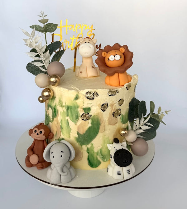 Safari-themed birthday cake with animal figurines including a lion, giraffe, elephant, monkey, and zebra, leafy decorations, and gold details.”