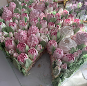 Square cake with pastel pink and white cream flowers including tulips and roses, with one slice removed.