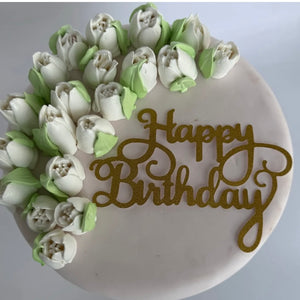 Birthday cake with white tulip-style cream flowers and a golden "Happy Birthday" topper on one side.