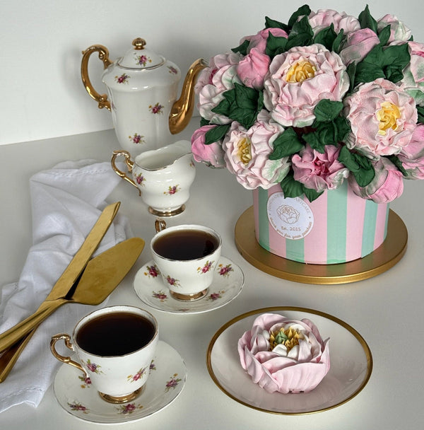 handmade marshmallow flower cake in a pastel striped box displayed with elegant vintage china teapot, teacups filled with tea, gold cake utensils, and a marshmallow flower on a dessert plate