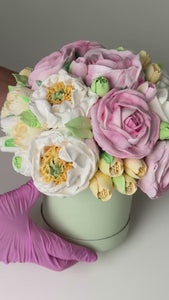 handmade marshmallow flower bouquet cake with pink, white and yellow blooms and green leaves arranged in a pastel green box, held in gloved hands